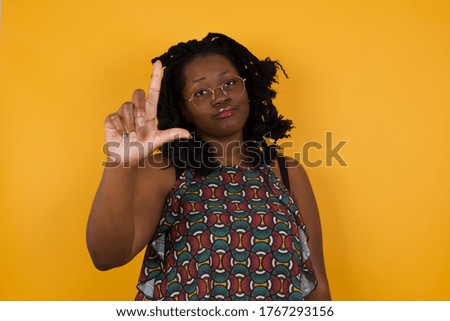 Young beautiful afro american woman wearing glasses over  yellow background making fun of people with fingers on forehead doing loser gesture mocking and insulting.