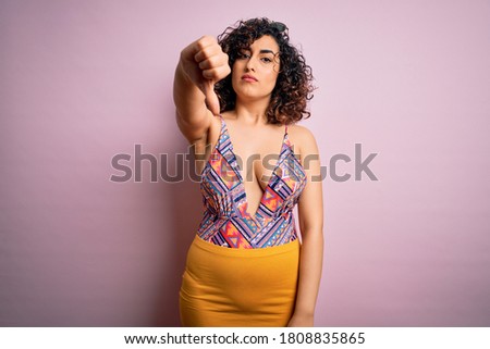 Young beautiful arab woman on vacation wearing swimsuit and sunglasses over pink background looking unhappy and angry showing rejection and negative with thumbs down gesture. Bad expression.