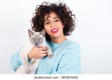 Young Beautiful Arab Woman With Short Curly Hair, Wearing Blue Knitted Sweater Standing Against White Background Holding And Hugging Her Siamese Cat Best Friend. 