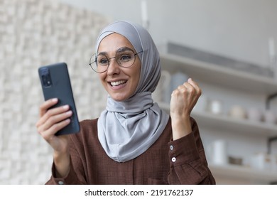 Young beautiful arab woman using phone smiling and celebrating success, muslim woman in hijab in living room holding smartphone - Powered by Shutterstock