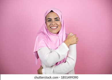 Young beautiful arab woman wearing islamic hijab over isolated pink background hugging oneself happy and positive, smiling confident