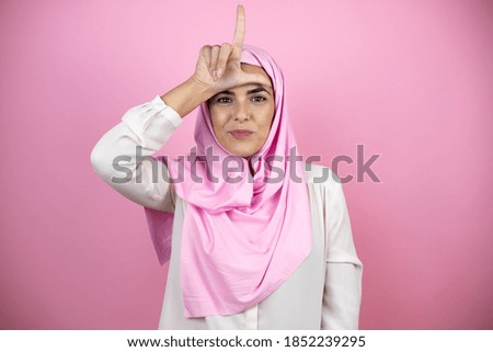 Young beautiful arab woman wearing islamic hijab over isolated pink background making fun of people with fingers on forehead doing loser gesture mocking and insulting.