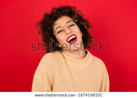 Young beautiful Arab woman wearing knitted sweater standing against red background with broad smile, shows white teeth, feeling confident rejoices having day off.
