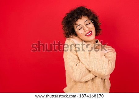 Young beautiful Arab woman wearing knitted sweater standing against red background. Hugging oneself happy and positive, smiling confident. Self love and self care.