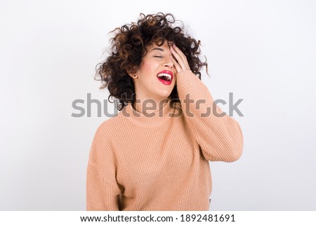 Young beautiful Arab woman wearing knitted sweater standing against white background makes face palm and smiles broadly, giggles positively hears funny joke poses