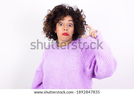 Young beautiful Arab woman wearing knitted sweater standing against white background purses lip and gestures with hand, shows something very little.