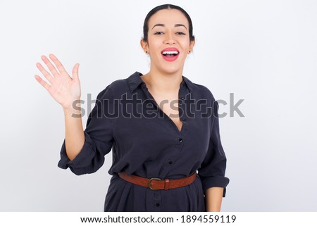 young beautiful Arab woman wearing gray dress against white studio background waiving saying hello or goodbye happy and smiling, friendly welcome gesture.