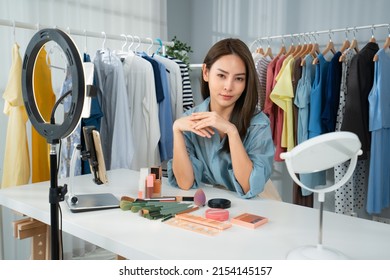 A Young Beautiful Asian Beauty Blogger Sitting At A Table With Cosmetics And Makeup Tools, Looking At Camera During Doing Makeup Tutorial Online Or Vlog In Front Of Mobiles And A Ring Light In A Room.