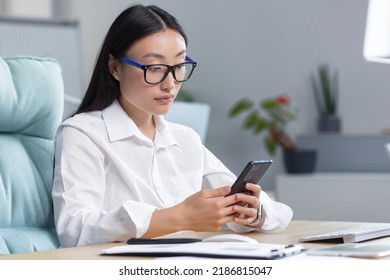 A Young Beautiful Asian Business Woman In Glasses Is Holding A Phone In Her Hands, Dialing A Message. Sitting At A Desk In A Modern Office.