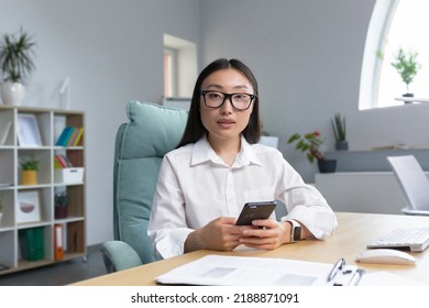 A Young Beautiful Asian Business Woman In Glasses Is Holding A Phone In Her Hands, Dialing A Message. Sitting At A Desk In A Modern Office, Looking At The Camera.