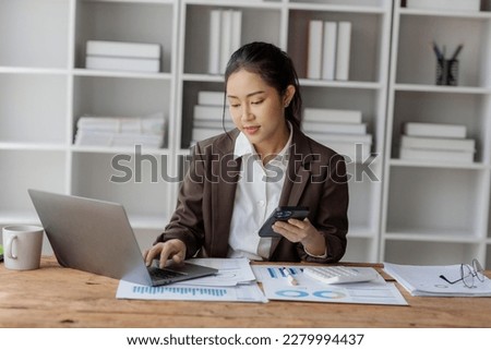 Young beautiful asian businesswoman using laptop and smart phone and calculator for doing financial mathematics on desk, tax, report, accounting, statistics and analytical research concept.