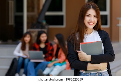 Young And Beautiful Asian College Student Girls Holding Books, Pose To Camera With Group Of Friends Blur In Background Against School Building. Learning And Friendship Of Teens Close Friend Concept.