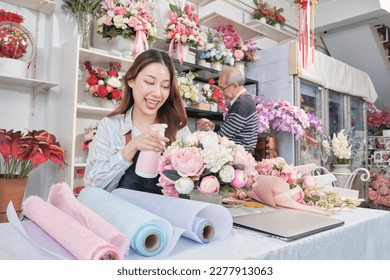 Young beautiful Asian female florist worker water spraying a flora bunch with old male employee, happy work in colorful flower shop store, fresh bloom bouquets decorating, SME business entrepreneur.  - Powered by Shutterstock