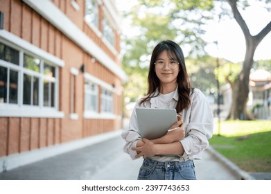 Young beautiful Asian female university student in casual clothes stands with her laptop computer in a campus garden. - Powered by Shutterstock