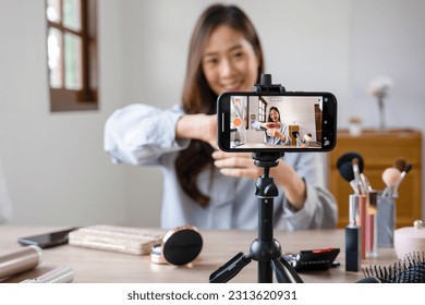 Young beautiful asian female vlogger, influencer sitting at table near window at home live streaming online from mobile phone while applying makeup as demonstration for viewers, selling cosmetics  - Powered by Shutterstock