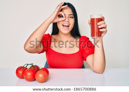 Young beautiful asian girl drinking glass of healthy tomato juice smiling happy doing ok sign with hand on eye looking through fingers 
