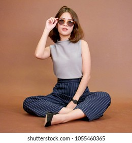 Young Beautiful Asian Girl Posting For Fashion Shoot. Sit On Ground. Touch Her Sunglasses. Wearing Gray Top, Blue Pants And Black Shoes