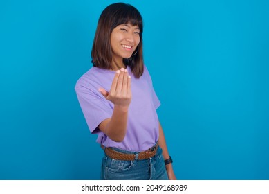 Young beautiful asian girl wearing purple t-shirt over blue background inviting to come with hand. Happy that you came