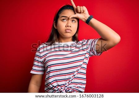 Young beautiful asian girl wearing casual striped t-shirt over isolated red background making fun of people with fingers on forehead doing loser gesture mocking and insulting.