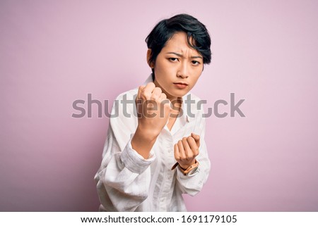 Young beautiful asian girl wearing casual shirt standing over isolated pink background Ready to fight with fist defense gesture, angry and upset face, afraid of problem