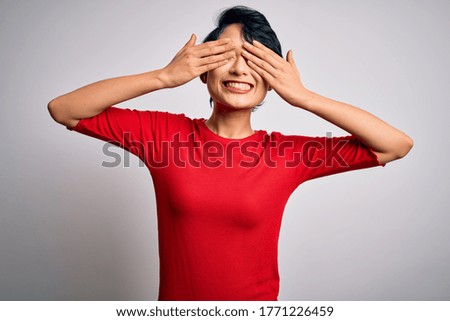 Young beautiful asian girl wearing casual red t-shirt standing over isolated white background covering eyes with hands smiling cheerful and funny. Blind concept.
