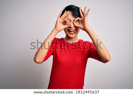 Young beautiful asian girl wearing casual red t-shirt standing over isolated white background doing ok gesture like binoculars sticking tongue out, eyes looking through fingers. Crazy expression.