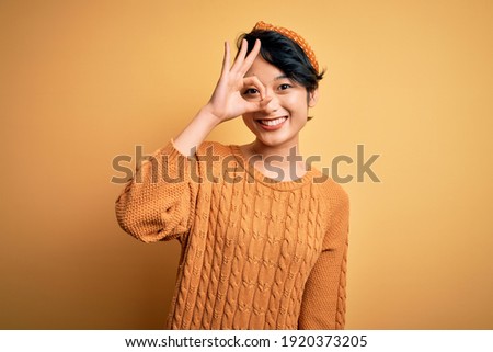 Young beautiful asian girl wearing casual sweater and diadem standing over yellow background doing ok gesture with hand smiling, eye looking through fingers with happy face.