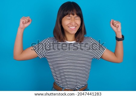 Young beautiful asian girl wearing striped t-shirt over blue background with surprised expression keeps hands under chin keeps lips folded makes funny grimace