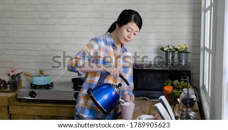 young beautiful asian japanese female filling mug with hot water while brewing beverage in kitchen. pretty elegant lady in shirt pour from kettle to cup making tea at home cooking place in morning.