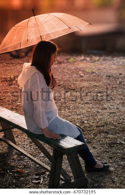 Young beautiful Asian lady wearing white rain\
coat, holding umbrella and sitting on a wooden bench with sad\
feeling or waiting or thinking of\
someone.