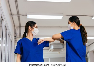 Young Beautiful Asian Medical Nurses In Scrub Uniform Wearing Face Mask Doing Elbow Bump At Hospital. Professional Medic At Workplace During Covid19 Pandemic. New Normal Greeting Concept