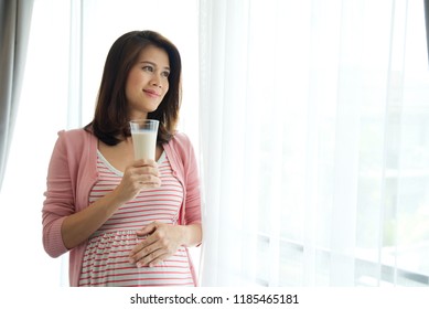 Young beautiful Asian pregnant woman standing in front of white curtain is holding milk on right hand and touching her tummy on left hand looking outside with smile. Pregnancy woman health concept.