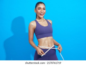 Young Beautiful Asian Sportswoman Holding Measuring Tape Around Her Waist Over Blue Background. Healthy And Fit Strong Female Concept.