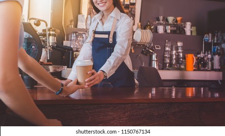 Young beautiful Asian woman barista wear blue apron holding hot coffee cup served to customer at bar counter in coffee shop with smile face.Concept of cafe and coffee shop small business.Vintage tone