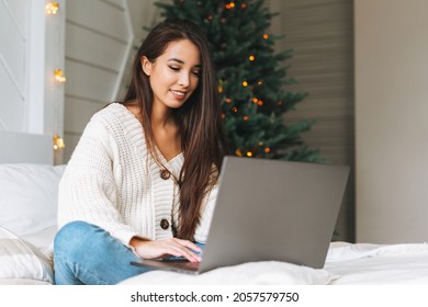 Young beautiful asian woman with dark long hair in cozy white knitted sweater using laptop on bed in room with Christmas tree. Portrait of carefree happy teenager girl student in house in christmas  - Powered by Shutterstock