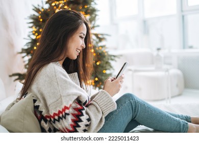 Young beautiful asian woman with dark long hair in cozy sweater using mobile smartphone on bed in room with Christmas tree 
