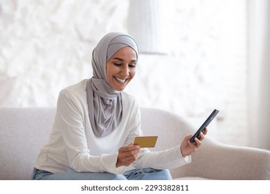 Young beautiful Asian woman in hijab sitting on sofa at home, Muslim woman holding bank credit card and phone in hand, happily shopping online in online store. - Powered by Shutterstock