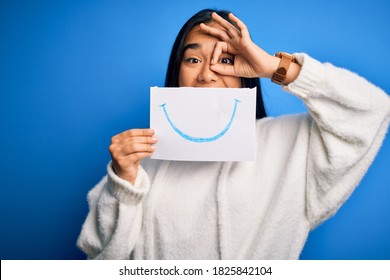Young beautiful asian woman holding paper and smile draw mouth over blue background and happy face smiling doing ok sign and hand eye looking through fingers