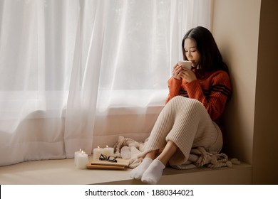 Young Beautiful Asian Woman Holding Cup Of Coffee And Marshmallows On Top, Sitting At Home And Looking Out The Window. Happy Girl Drinking Chocolate In Sweater In Cold Weather Winter