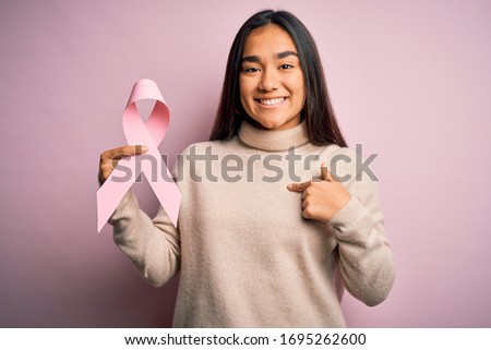 Young beautiful asian woman holding pink cancer ribbon symbol over isolated background with surprise face pointing finger to himself