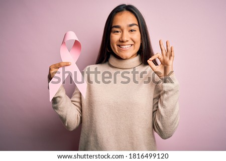 Young beautiful asian woman holding pink cancer ribbon symbol over isolated background doing ok sign with fingers, excellent symbol