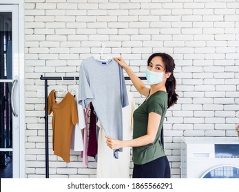 Young Beautiful Asian Woman, Housewife In Casual Wearing Protective Face Mask Hanging Dry Shirt With Hanger On Clothesline After Washing Near Wash Machine In Laundry Room On White Wall Background.