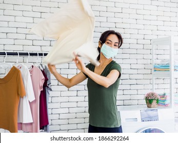 Young Beautiful Asian Woman, Housewife Wearing Casual Cloth And Protective Face Mask Shaking And Drying Shirt After Washing Before Hanging On Clothesline Near Wash Machine On White Wall Background.