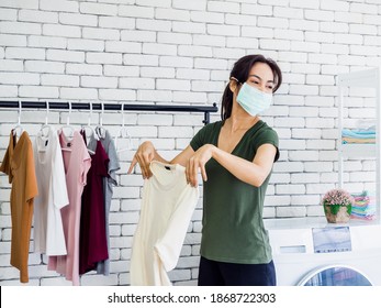 Young Beautiful Asian Woman, Housewife Wearing Casual Cloth And Protective Face Mask Shaking And Drying Shirt After Washing Before Hanging On Clothesline Near Wash Machine On White Wall Background.