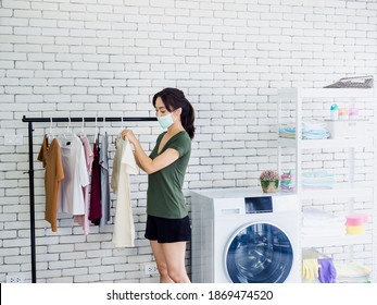 Young Beautiful Asian Woman, Housewife In Casual Wearing Protective Face Mask Hanging Dry Shirt With Hanger On Clothesline After Washing Near Wash Machine In Laundry Room On White Wall Background.