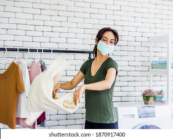 Young Beautiful Asian Woman, Housewife Wearing Casual Cloth And Protective Face Mask Shaking And Drying Shirt After Washing Before Hanging On Clothesline Near Wash Machine On White Wall Background.