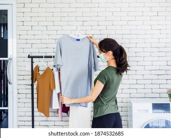 Young Beautiful Asian Woman, Housewife In Casual Wearing Protective Face Mask Hanging Dry Shirt With Hanger On Clothesline After Washing Near Wash Machine In Laundry Room On White Wall Background.