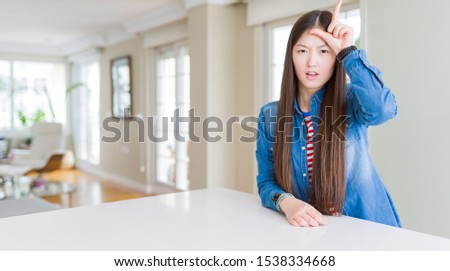 Young beautiful asian woman with long hair wearing denim jacket making fun of people with fingers on forehead doing loser gesture mocking and insulting.