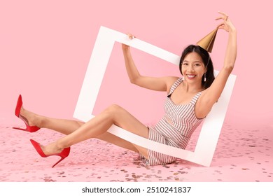 Young beautiful Asian woman in party hat with frame and confetti sitting on pink background - Powered by Shutterstock