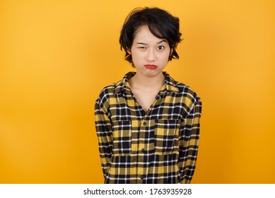 Young beautiful Asian woman with short hair wearing casual plaid shirt over yellow background making grimace and crazy face, screaming out of control, funny lunatic expressing freedom and wild.